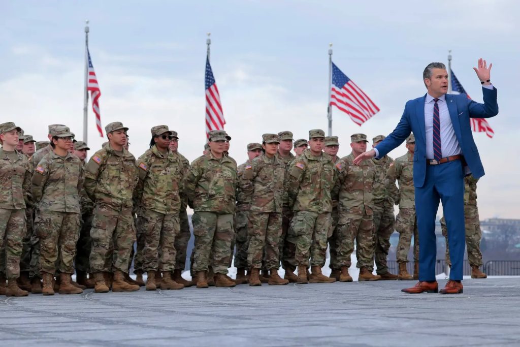 Man in suit addresses National Guard soldiers in formation with US flags in background.